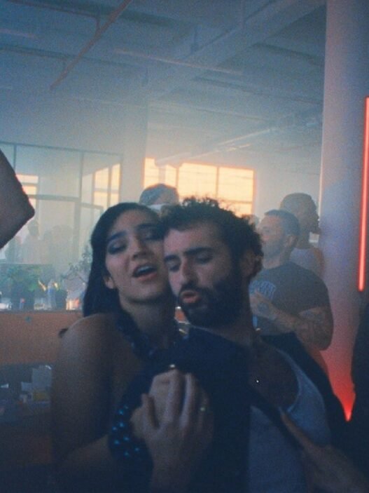 Una fiesta nocturna en un loft con humo y luz azul, con dos personas bailando abrazadas en primer plano