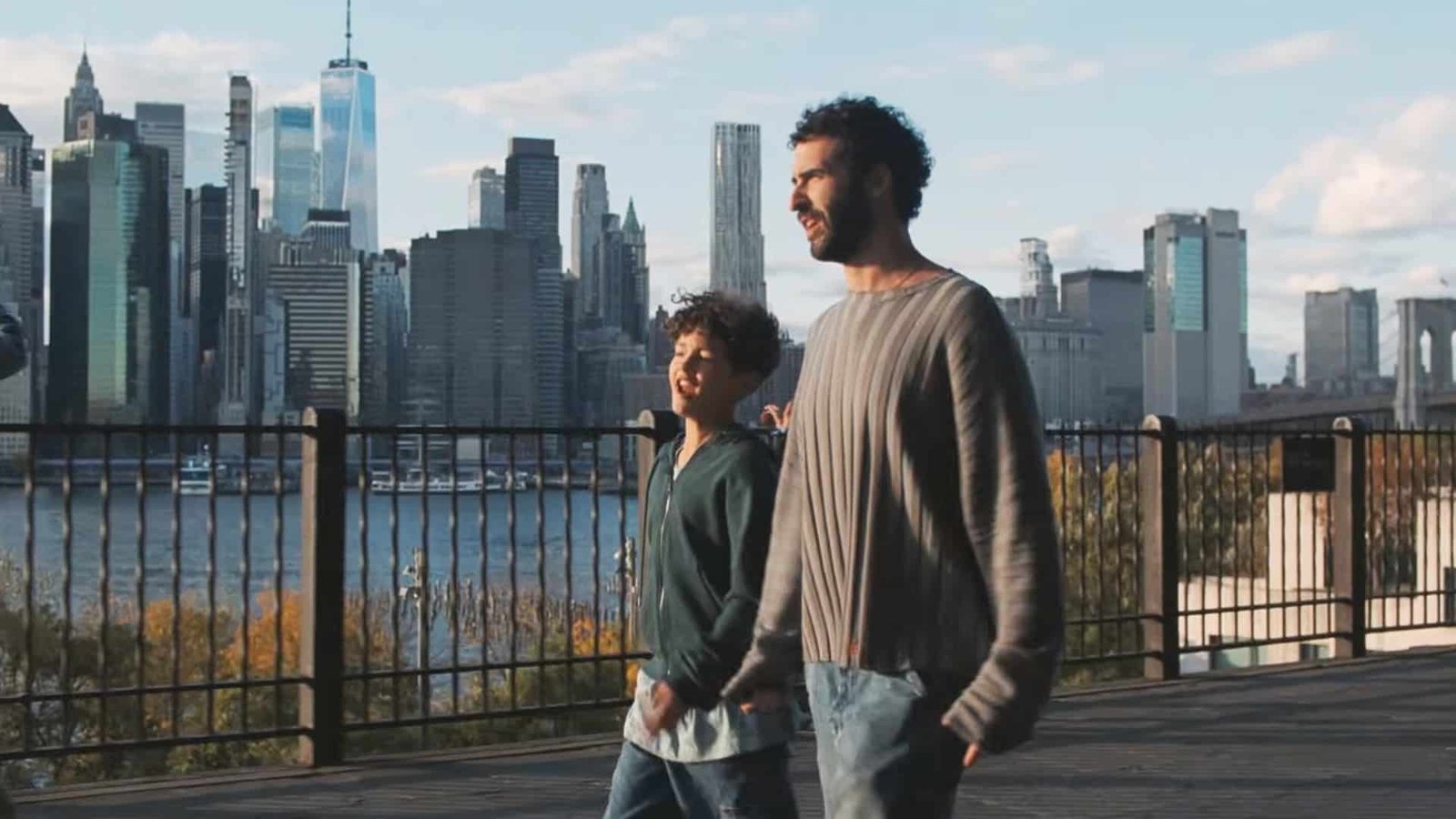 Un hombre y un niño caminan junto a una reja con el horizonte de Manhattan al fondo