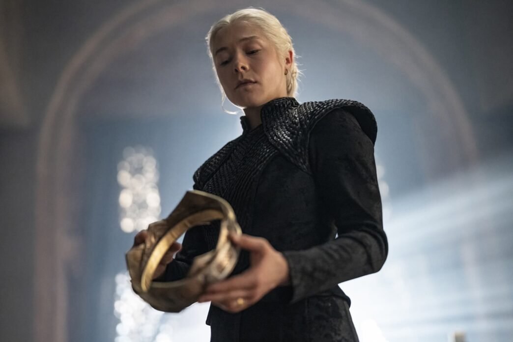 Rhaenyra Targaryen, vestida de negro, sosteniendo una corona dorada en un salón de piedra