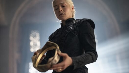 Rhaenyra Targaryen, vestida de negro, sosteniendo una corona dorada en un salón de piedra