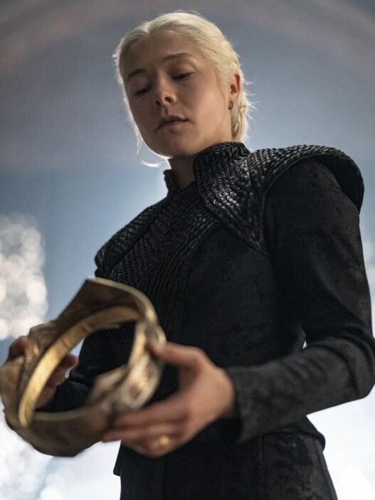 Rhaenyra Targaryen, vestida de negro, sosteniendo una corona dorada en un salón de piedra