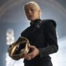 Rhaenyra Targaryen, vestida de negro, sosteniendo una corona dorada en un salón de piedra