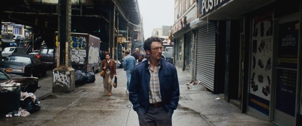 Irwin camina con las manos en los bolsillos por una calle de Nueva York bajo un tren elevado, rodeado de carros y graffiti de los años 80