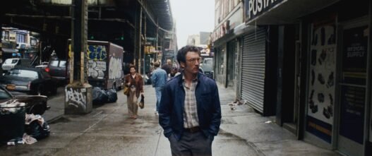 Irwin camina con las manos en los bolsillos por una calle de Nueva York bajo un tren elevado, rodeado de carros y graffiti de los años 80