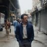 Irwin camina con las manos en los bolsillos por una calle de Nueva York bajo un tren elevado, rodeado de carros y graffiti de los años 80