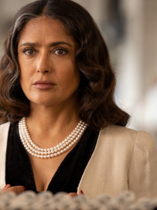 Salma Hayek como Nina, con collar de perlas y saco claro, mira de frente con gesto serio
