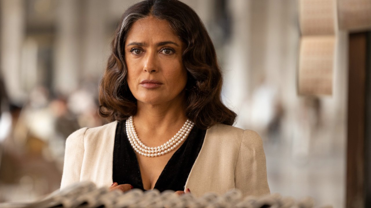 Salma Hayek como Nina, con collar de perlas y saco claro, mira de frente con gesto serio